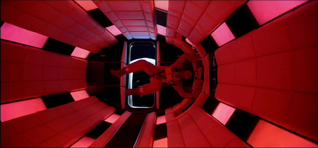2001: A Space Odyssey (1968) movie still: fullbody — A long cylindrical corridor is lined with padded red wall panels and glowing lights. A man in a…
