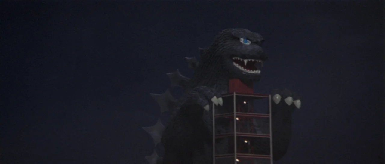 Godzilla Vs Gigan (1972) movie still: medium — A dark night setting features a large monster standing behind a metal tower structure. The creature's…