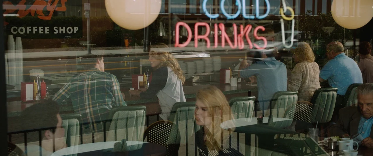 Happy Death Day (2017) movie still: wide — A view through a storefront window shows people eating inside a classic American eatery. Red neon text…
