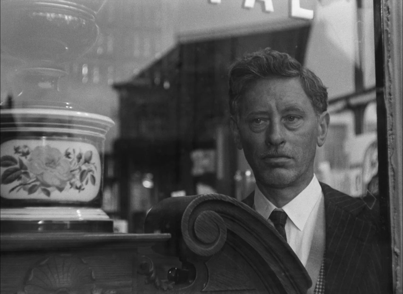 Hiroshima Mon Amour (1959) movie still: closeup — A man looks through the glass of a store display. The frame includes a decorative porcelain vase…