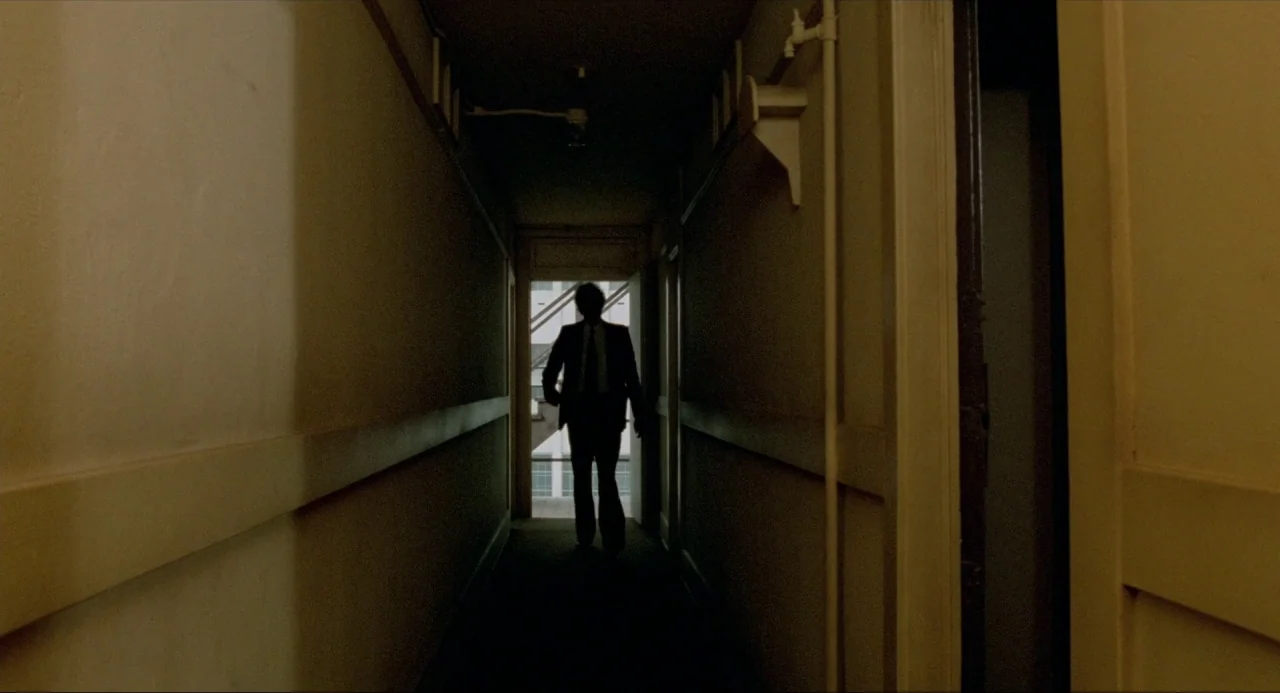 The Driver (1978) movie still: wide — A dark, narrow building corridor is flanked by plain beige walls and a doorway. A man in a suit walks toward…