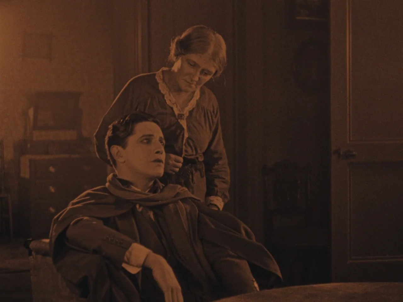 The Lodger (1927) movie still: medium — A dimly lit room features a man sitting while a woman stands behind him. The environment is sparsely…
