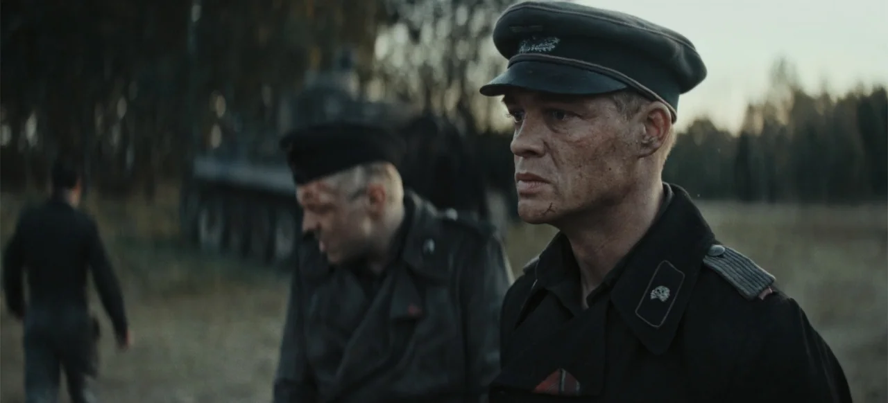 The Tank (2025) movie still: medium — An open field at dusk with a parked military tank in the background. Two soldiers stand in the foreground…