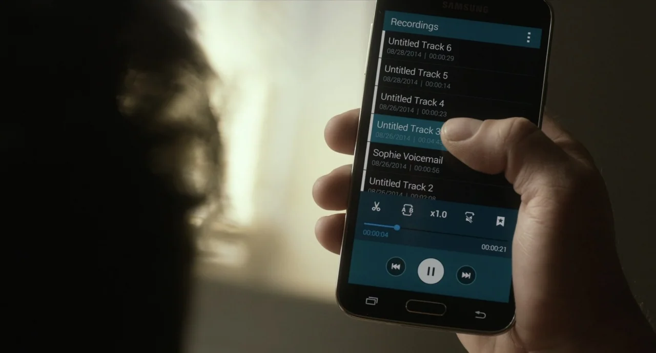 We Are Your Friends (2015) movie still: closeup — A person holds a black smartphone displaying a digital recording list. The out-of-focus subject in…