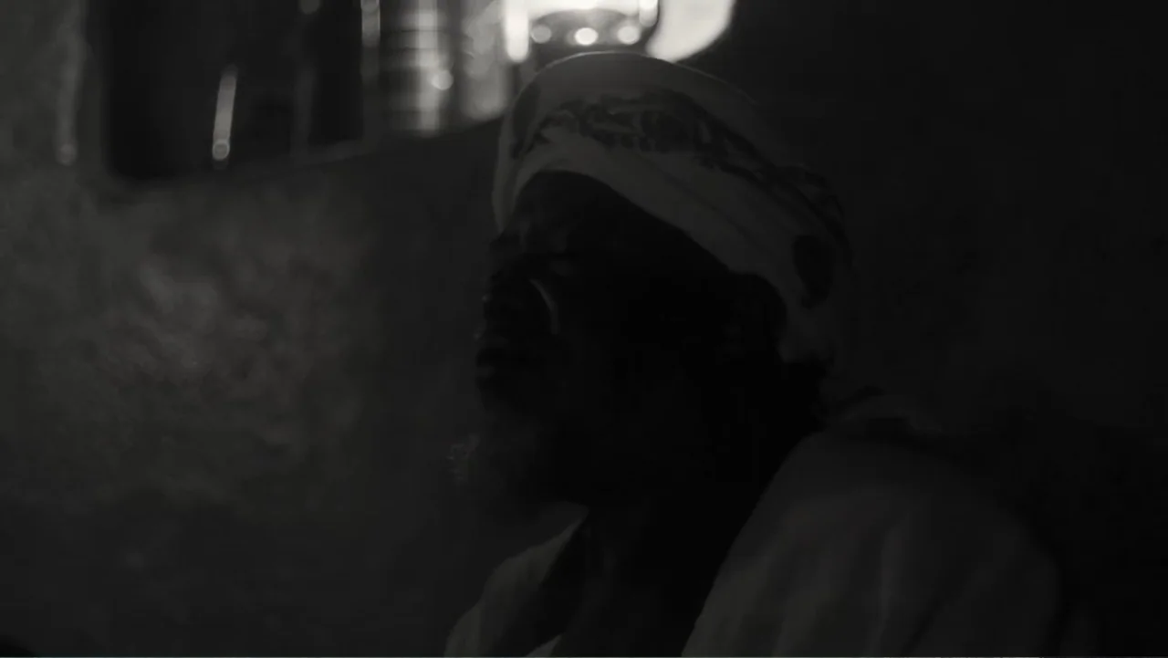 Faya Dayi (2021) movie still: closeup — A dark interior space is lit by a single glowing practical lamp in the background. An elderly man with a…