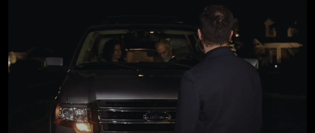 Ordinary Things (2001) movie still: medium — A dark residential driveway features a parked grey SUV at night. A man in a dark blazer stands in the…