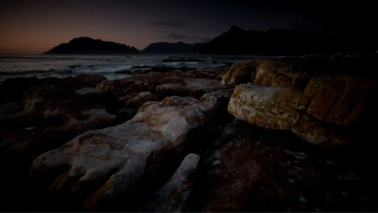 Air Jaws: Night Stalker (2016) movie still: establishing — A dark rocky shoreline meets a calm sea at the edge of the horizon. Waves wash over the…