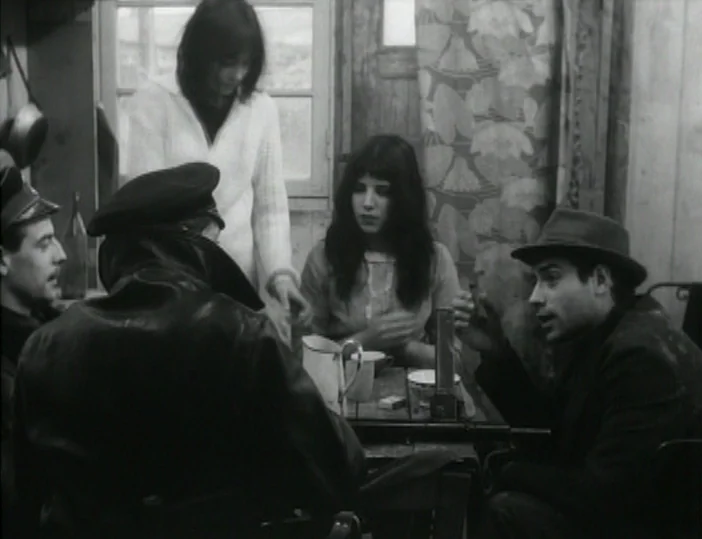 Les Carabiniers (1963) movie still: medium — A small interior space with floral wallpaper and a window in the background. A group of people gather…