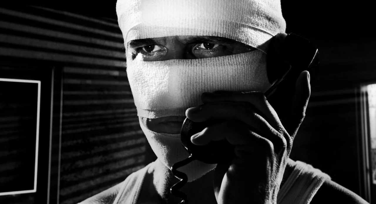 Sin City: A Dame to Kill For (2014) movie still: closeup — A dark interior with a man whose head is heavily swaddled in white bandages. The man…