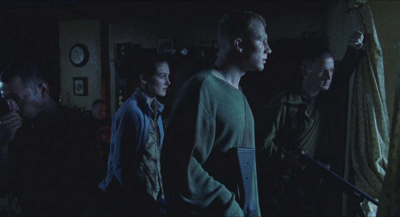 Dog Soldiers (2002) movie still: medium — A dark, dimly lit domestic space filled with shadows and muted tones. Several individuals stand grouped…
