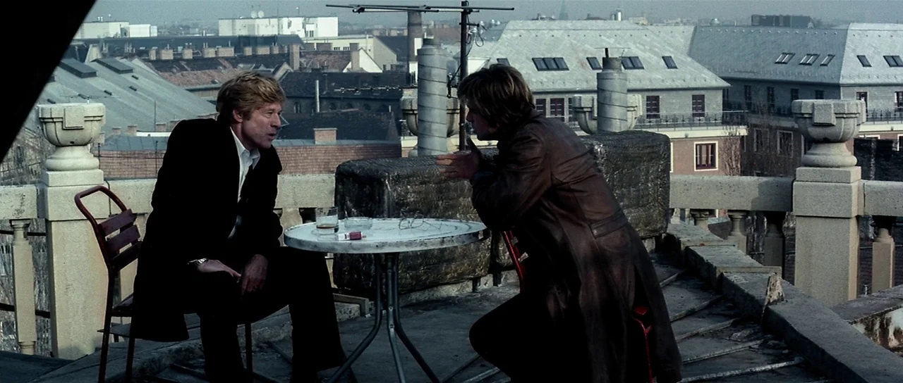 Spy Game (2001) movie still: medium — An urban rooftop with decorative stone pedestals and a view of gray city buildings. Two men sit across from…