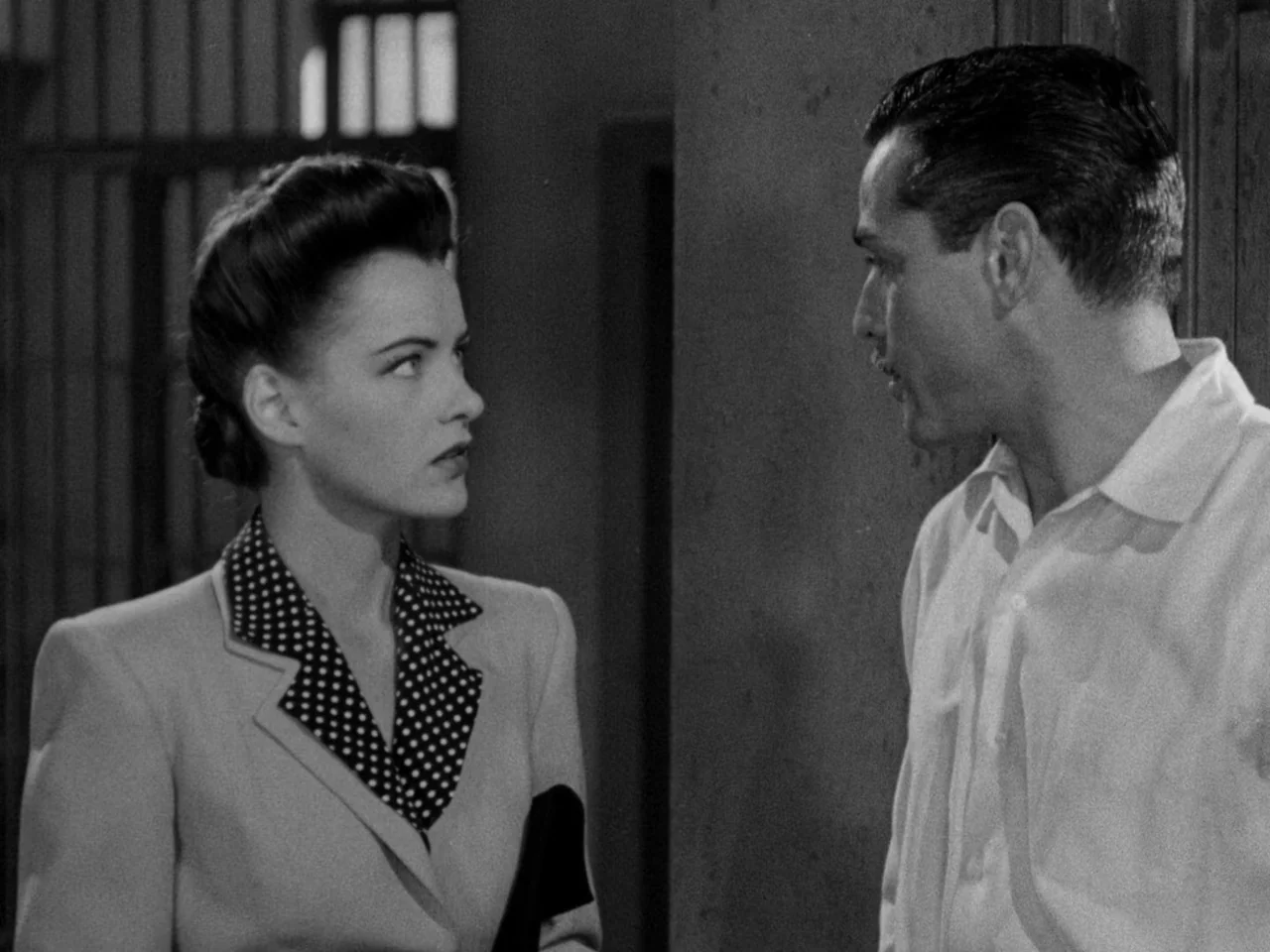 Phantom Lady (1944) movie still: medium — A stark interior setting defined by vertical metal bars in the background. A woman and man stand facing…