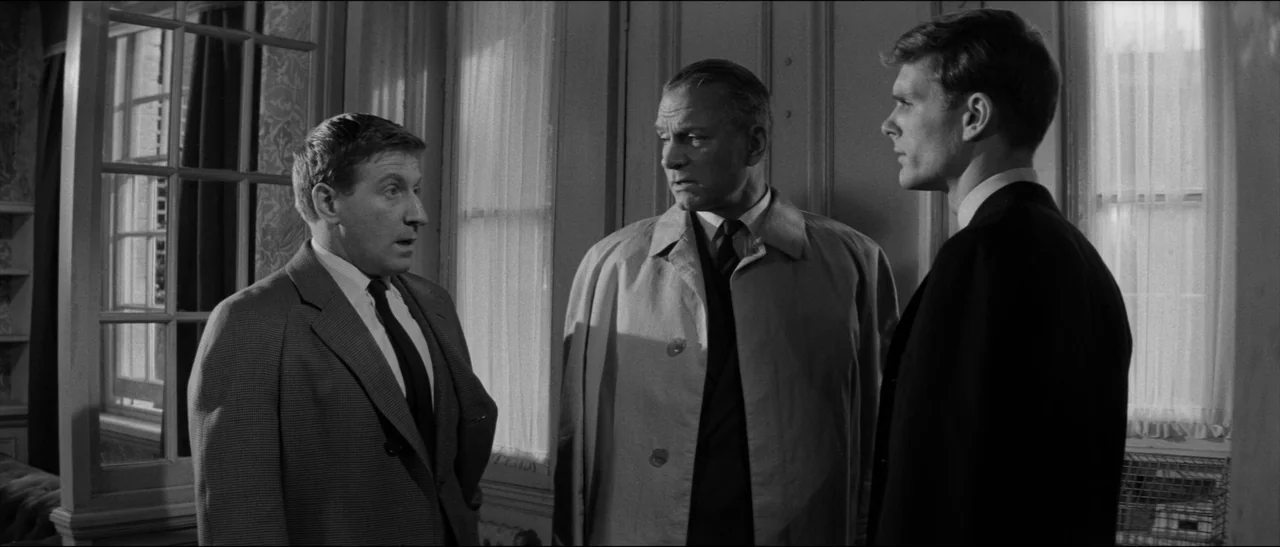 Bunny Lake Is Missing (1965) movie still: medium — Three men stand together in a dimly lit study with large window panes. They are engaged in a…