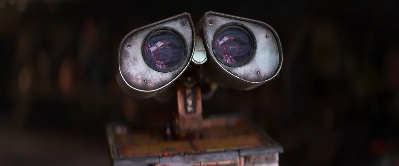 Wall-E (2008) movie still: closeup — A dark, blurred space contains a weathered, rusty robotic head with circular eyes. The robot is facing the…