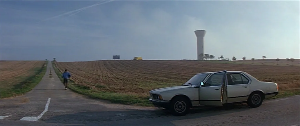 IP5 (1992) movie still: wide — A flat, expansive rural landscape features plowed fields and a tall, thin water tower. A man in sportswear jogs away…