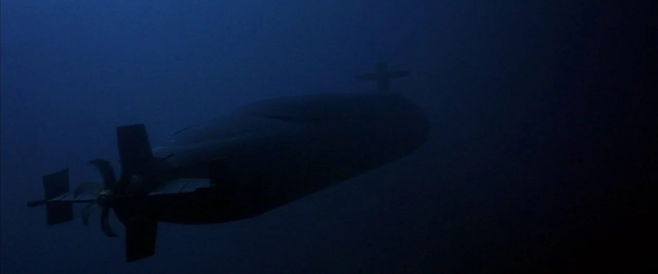 Crimson Tide (1995) movie still: wide — A dark, massive military submarine moves through the deep, murky blue underwater abyss. The vessel is…