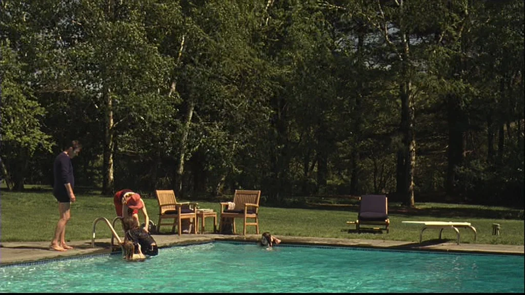 Margot At The Wedding (2007) movie still: wide — An outdoor swimming pool is surrounded by green grass and tall leafy trees. A man stands at the…