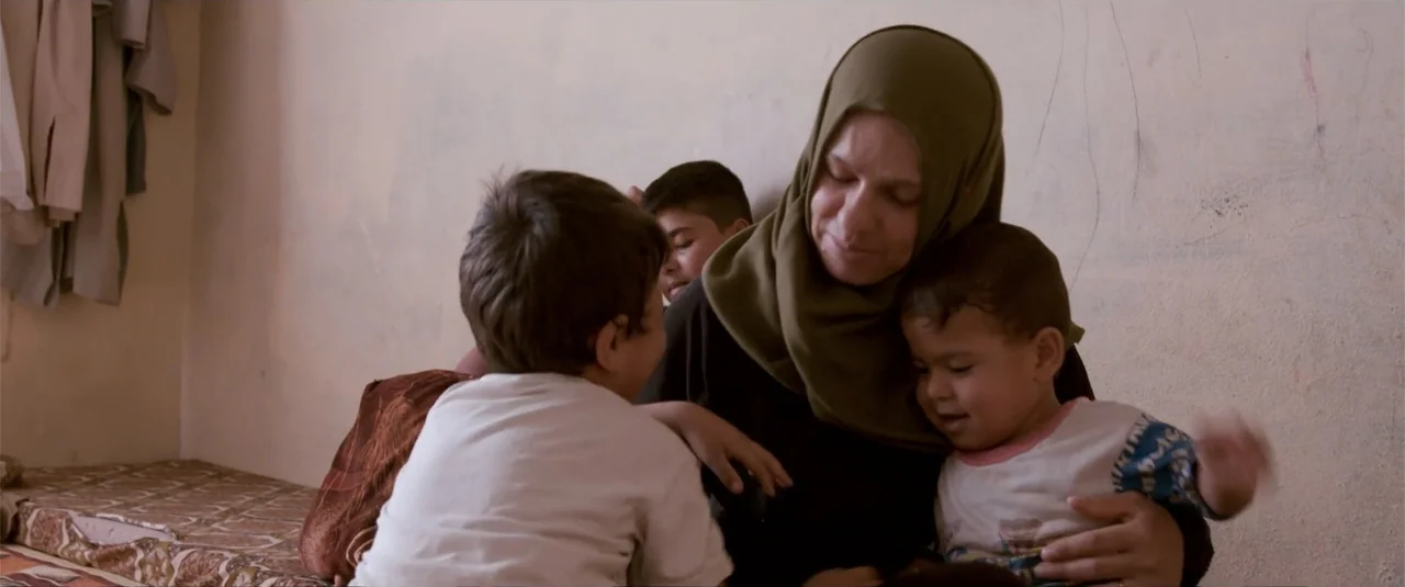 Simple as Water (2021) movie still: medium — A sparsely furnished room with a plain, light-colored wall and a mattress on the floor. A woman in a…