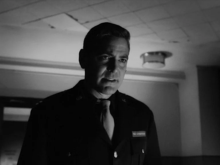 The Good German movie still: medium — A dimly lit indoor corridor with a drop-tile ceiling and industrial lighting. A man in a dark military-style…