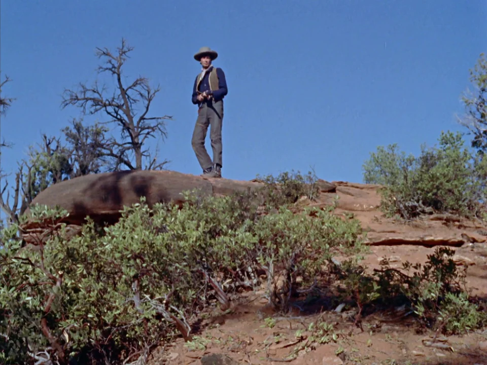 Johnny Guitar (1954) movie still: fullbody — A man stands atop a sandstone rock formation in a vast, arid desert landscape. He holds a rifle and…
