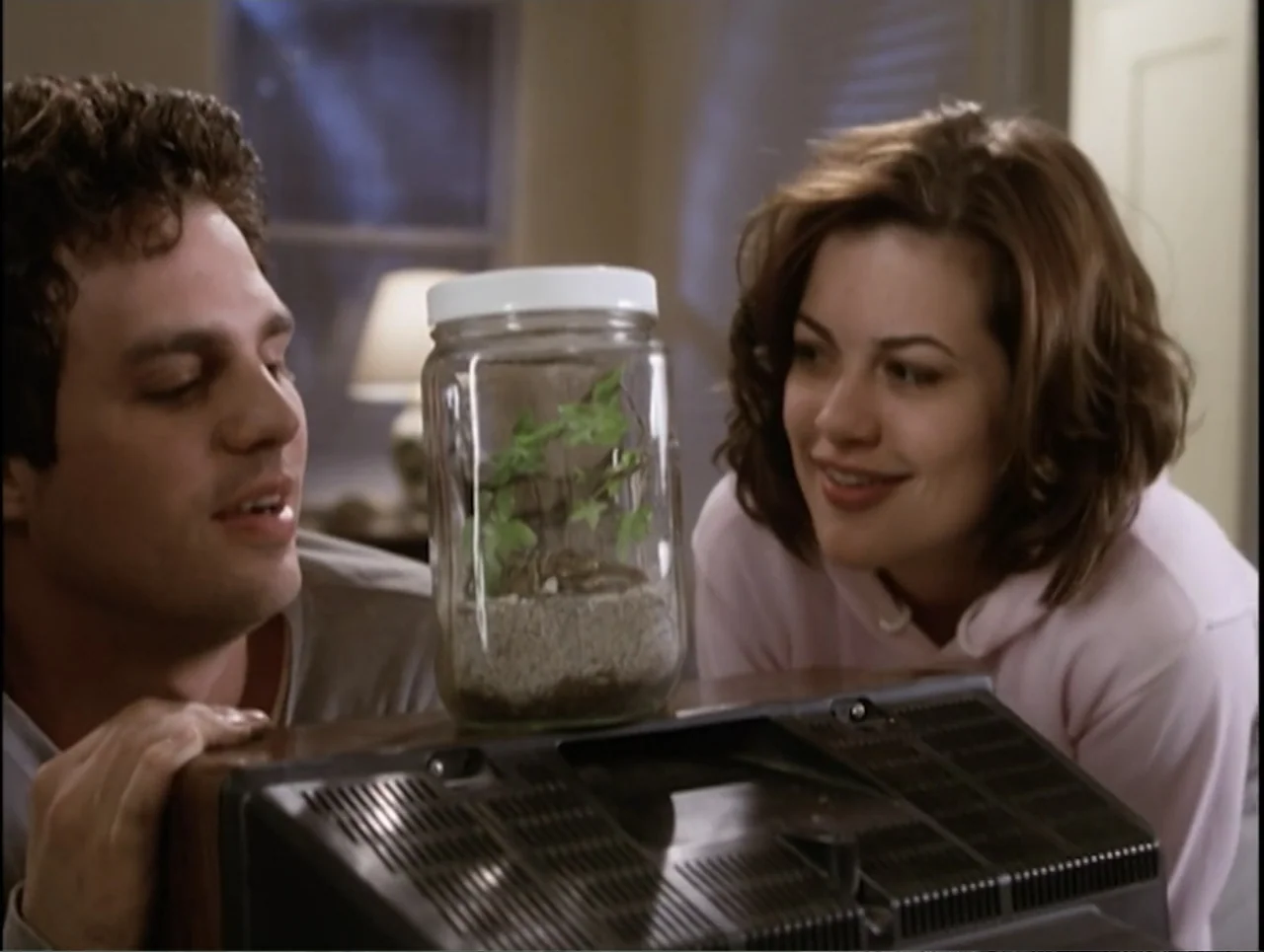 Apartment 12 (2001) movie still: medium — A domestic space features a man and a woman leaning over a television console. A glass jar containing a…