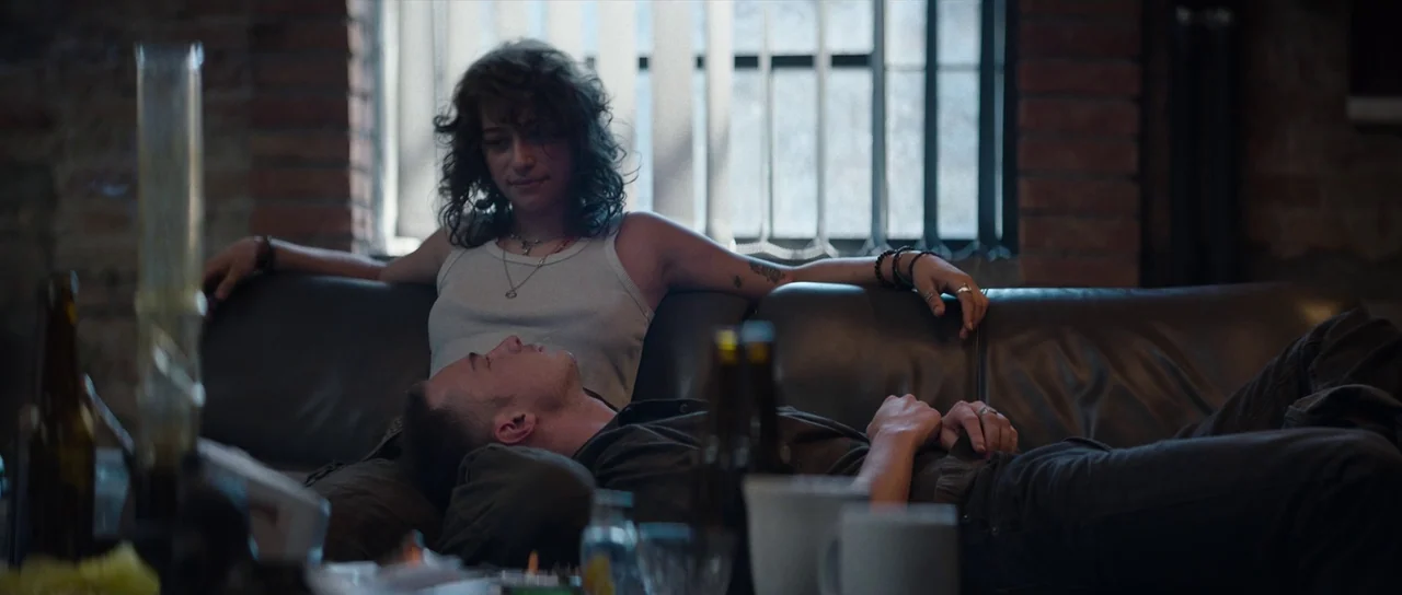 Hellraiser (2022) movie still: medium — An urban industrial apartment space with exposed brick walls and a slatted window. A woman sits on a leather…