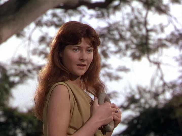 The River (1951) movie still: medium — A woman stands beneath a large tree during the day. She holds a smooth stone object against her chest with…