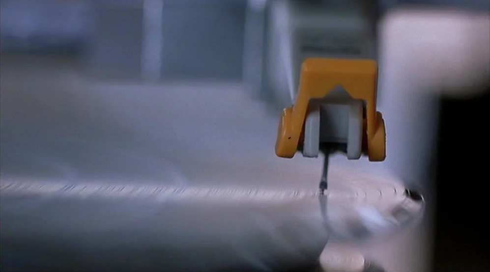 Jackie Brown movie still: closeup — A close-up view shows the orange-trimmed plastic arm of a specialized recording device. The stylus rests within…