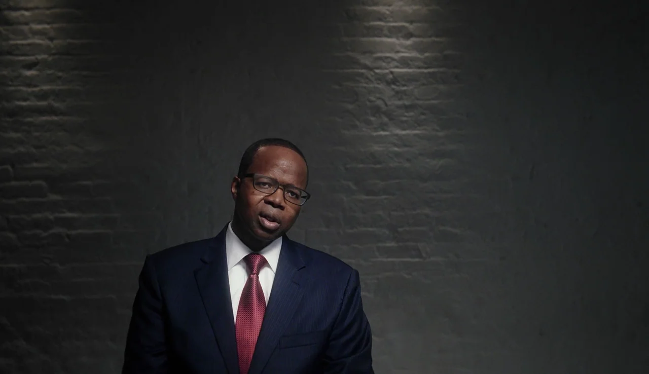 13th (2016) movie still: medium — A man in a suit stands against a dark grey painted brick wall. He is positioned in front of the wall while looking…