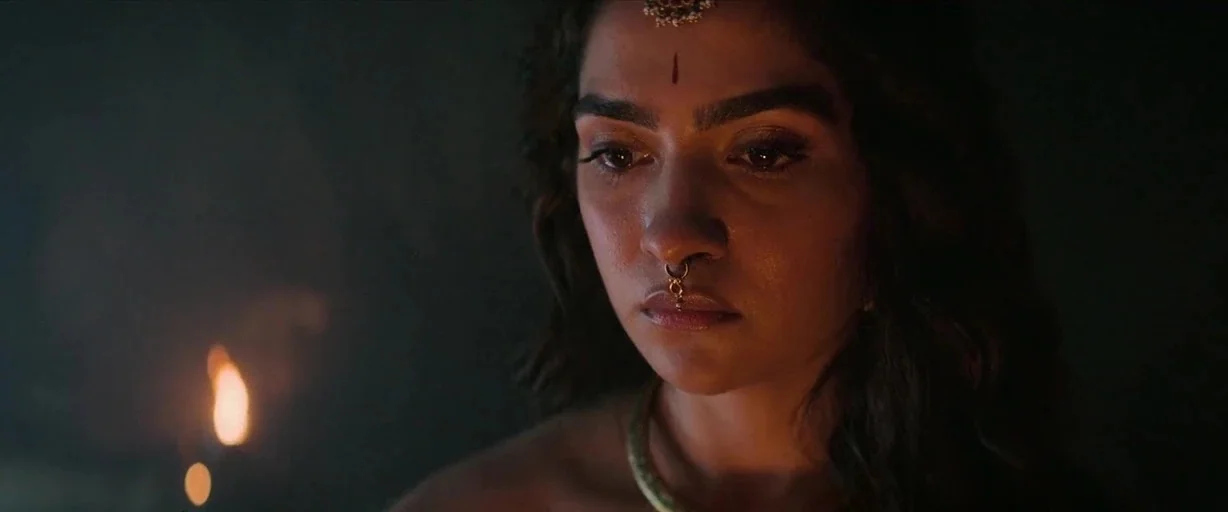 Kantara: A Legend - Chapter 1 (2025) movie still: closeup — A dimly lit interior space is cast in shadows. A young woman with traditional jewelry…