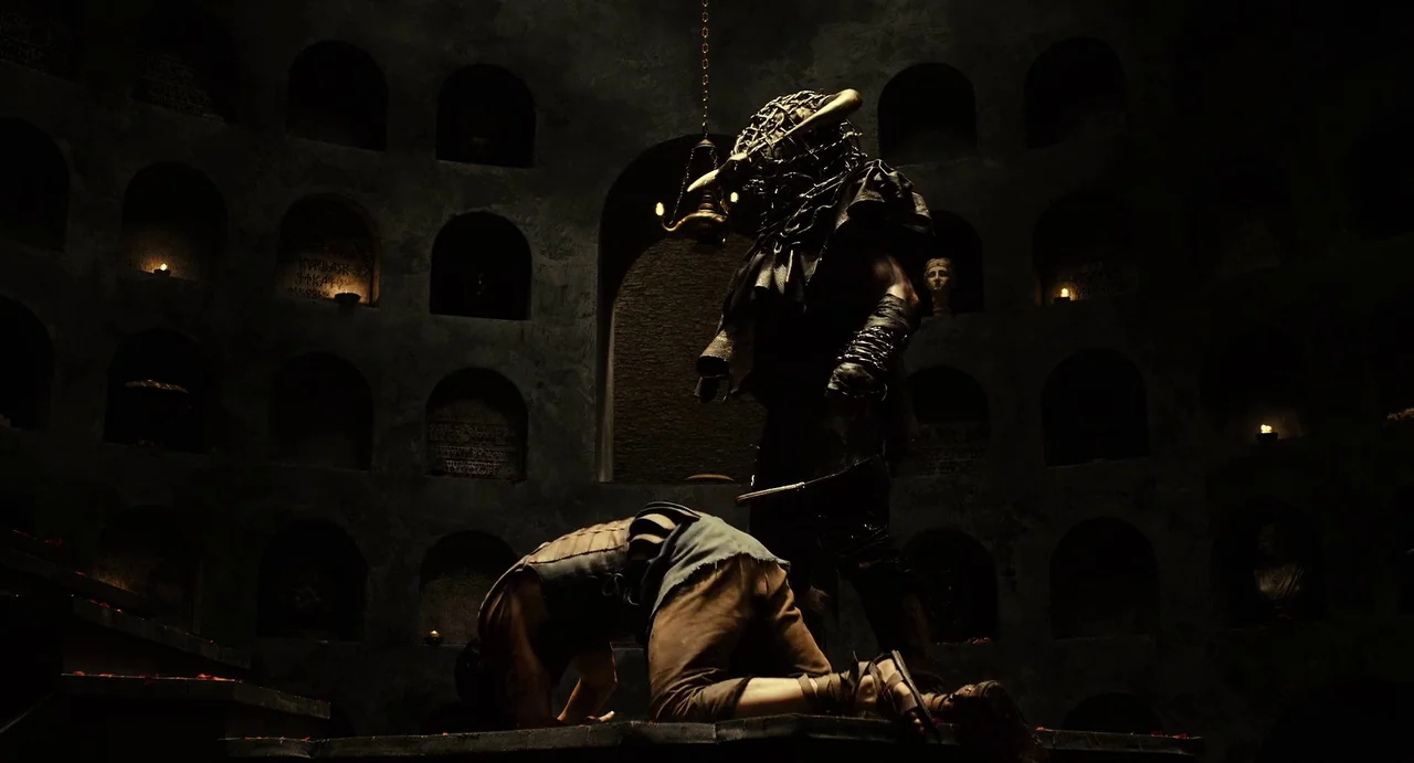 The Immortals (2011) movie still: fullbody — A dark, circular stone burial chamber features arched niches containing small flickering candles. An…