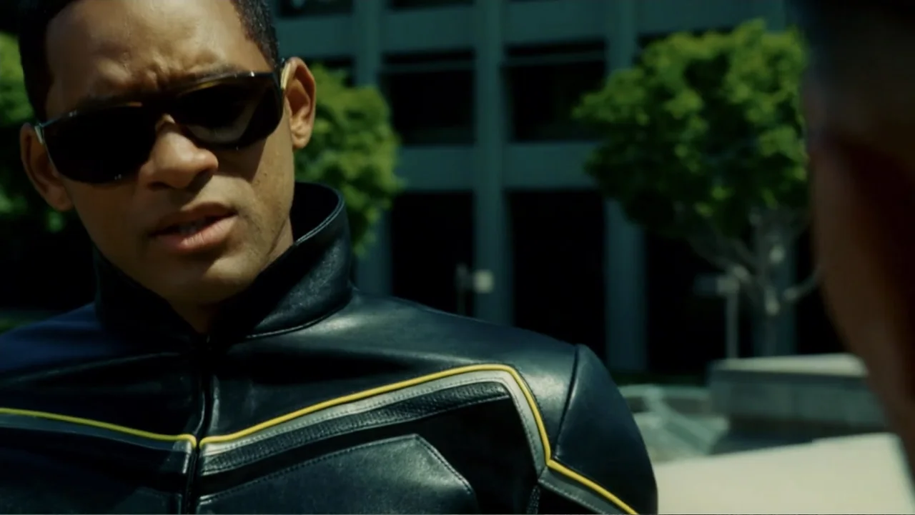 Hancock (2008) movie still: medium — A paved urban plaza features a blurred office building and green tree in background. A man in a leather jacket…