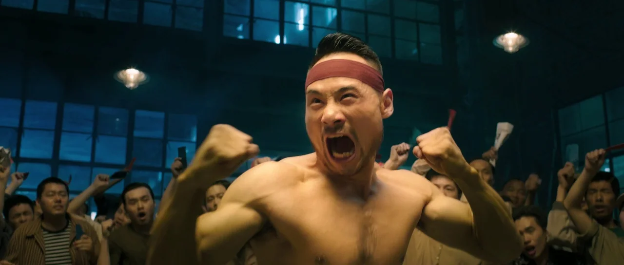 Ip Man 3 (2015) movie still: medium — A crowded industrial space with large paned glass windows and low hanging lights. A bare-chested man shouts…