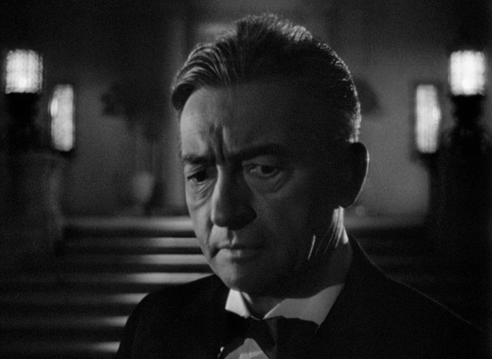 Notorious (1946) movie still: closeup — A dimly lit interior space features two glowing lamps positioned in the distant background. A middle-aged…