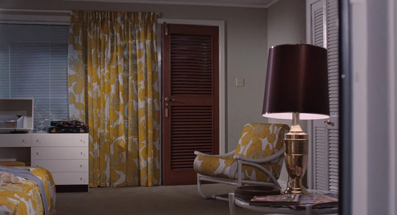 Live and Let Die (1973) movie still: wide — A mid-century motel room features yellow floral curtains and matching fabric on a chair. A white chest…