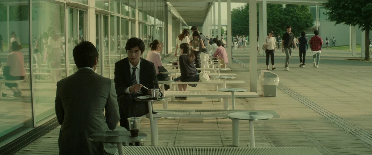 Creepy (2016) movie still: medium — An outdoor paved walkway with modern white architecture and glass walls lined with seating. Two men in suits sit…