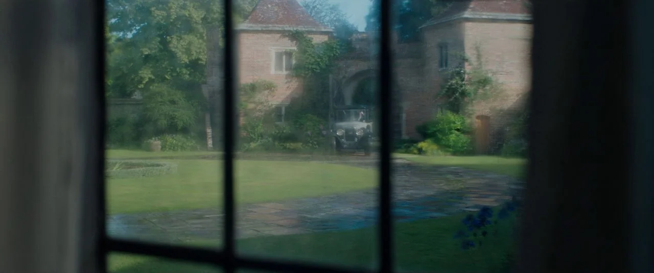 Rebecca movie still: establishing — A stone manor with ivy-covered walls stands at the end of a stone path. A vintage car is parked near the arched…