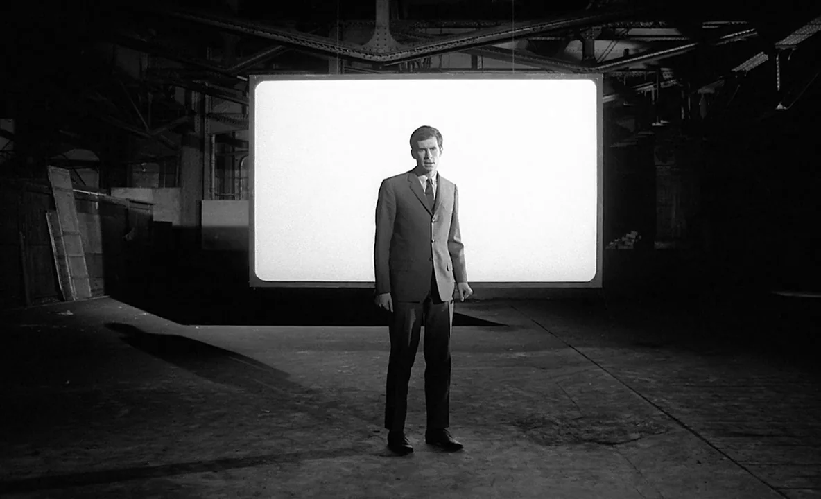 The Trial (1962) movie still: fullbody — A man stands in a dark, cavernous industrial space with exposed metal beams above him. A large, illuminated…