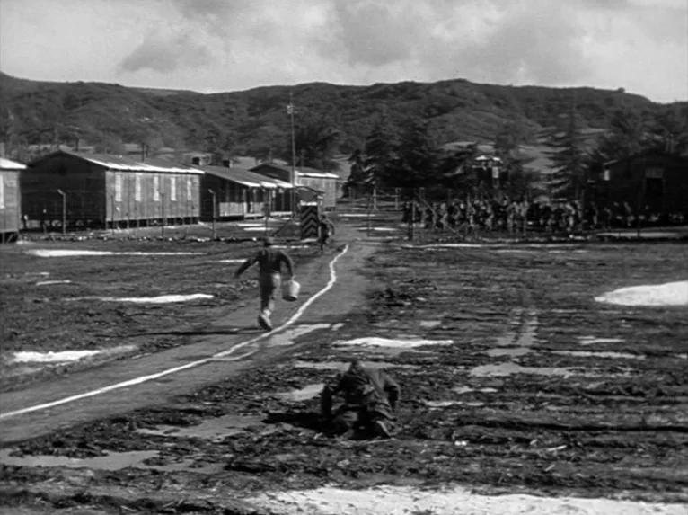 Stalag 17 (1953) movie still: wide — A POW camp with wooden barracks aligned along a muddy central path. One man runs down the path while another…