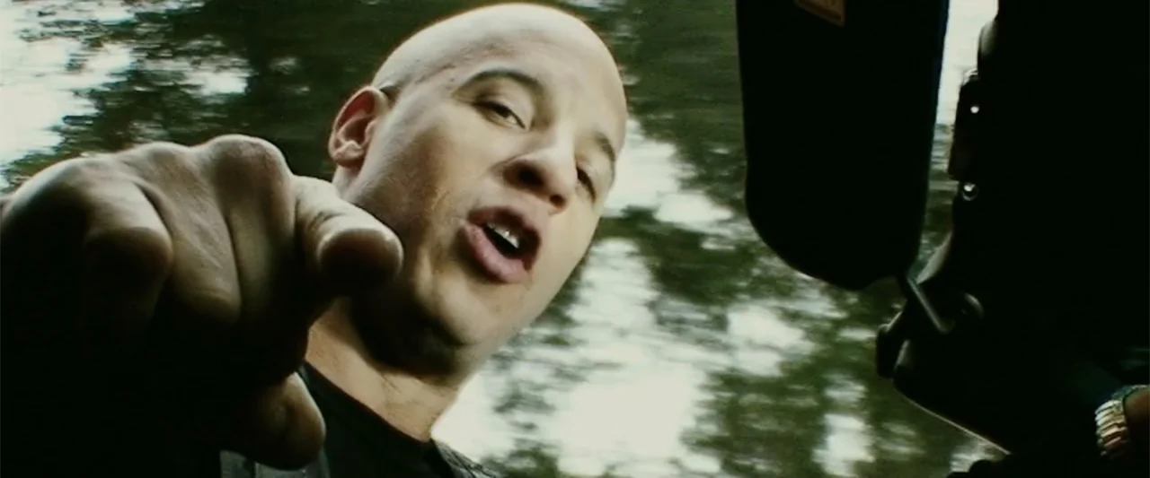 xXx (2002) movie still: closeup — A bald man sits inside a moving vehicle with blurred trees visible outside. He points his finger toward the viewer…