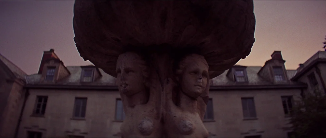 Braid (2018) movie still: closeup — A stone fountain with carved female figures stands in front of a colonial building. The sky is a gradient of…