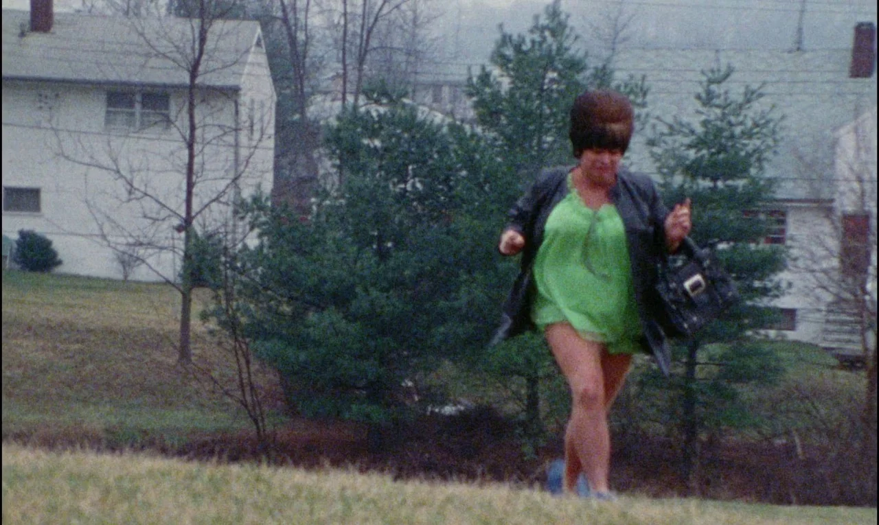 Female Trouble (1974) movie still: fullbody — A suburban residential area with houses and evergreen trees in the background. A woman in a green…