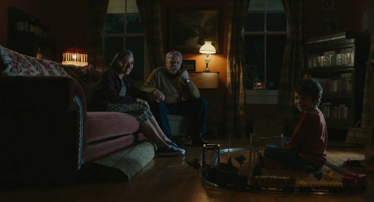 The Life of Chuck (2024) movie still: wide — A dimly lit room features a sofa and bookshelves in the evening. A young boy plays with a model train…