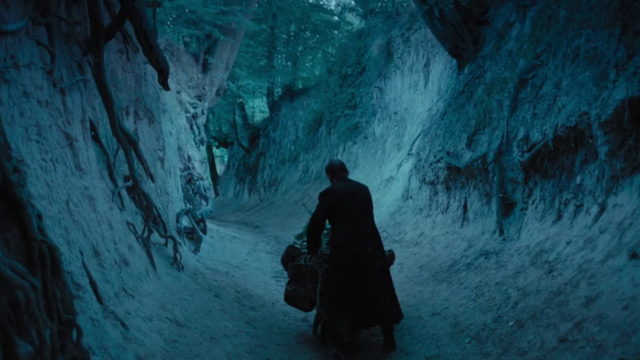 The Field Guide to Evil (2018) movie still: wide — A narrow dirt path is flanked by steep eroded earth banks with exposed roots. An elderly man in a…