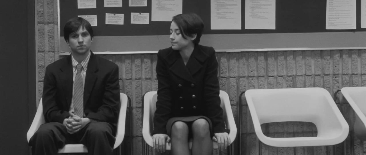 Polytechnique (2009) movie still: medium — A man and a woman sit in white chairs against a brick wall. Several documents are pinned to a bulletin…