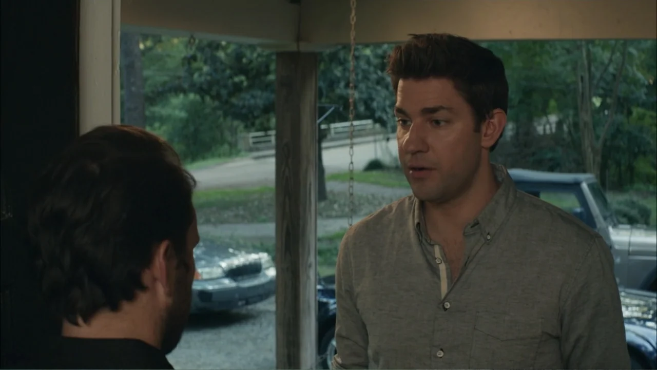 The Hollars (2016) movie still: medium — A residential garage doorway looks out onto a driveway with parked vehicles. A man in a grey shirt engages…