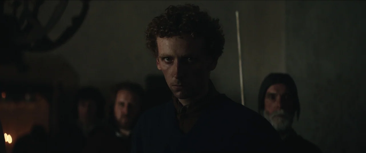 The King movie still: medium — A dimly lit stone chamber with rough walls houses a small group of figures. The central young man faces forward while…