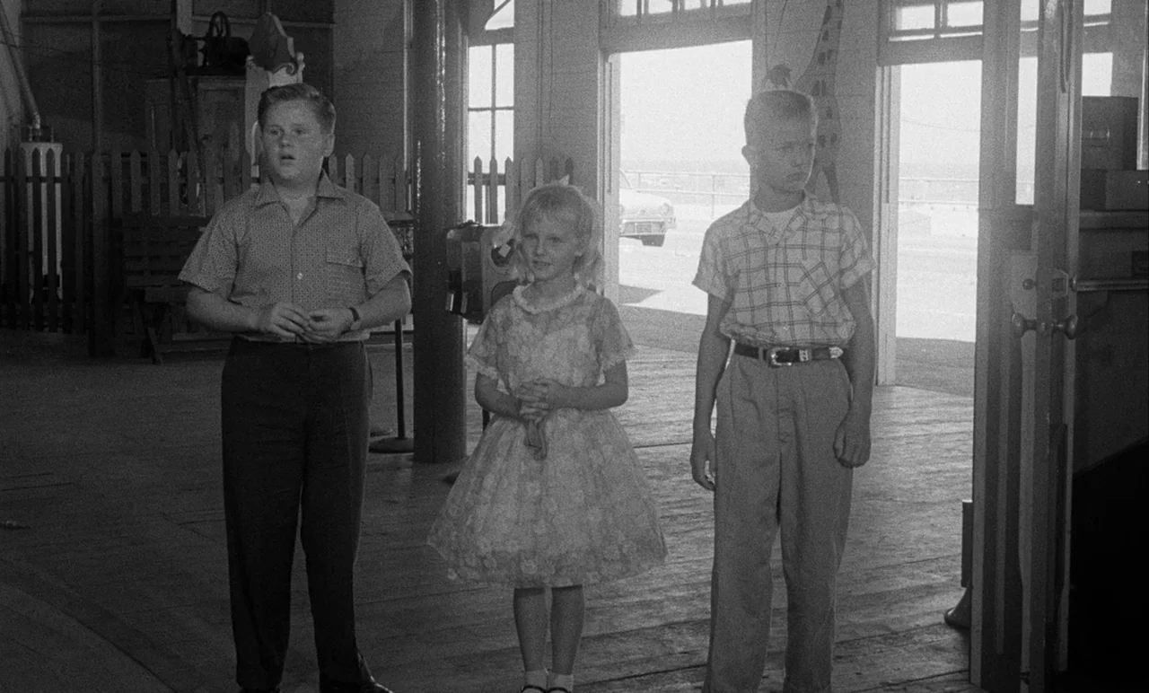 Night Tide (1961) movie still: fullbody — An interior waiting area with wooden floors and open doorways to the outdoors. Three children stand…