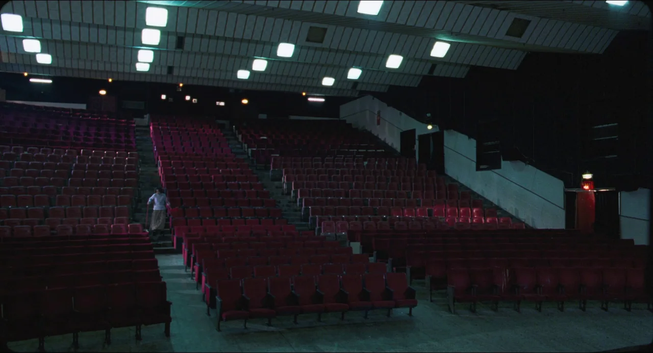 Goodbye, Dragon Inn (2003) movie still: wide — A large, empty cinema hall features rows of red chairs under bright fluorescent lights. A lone person…