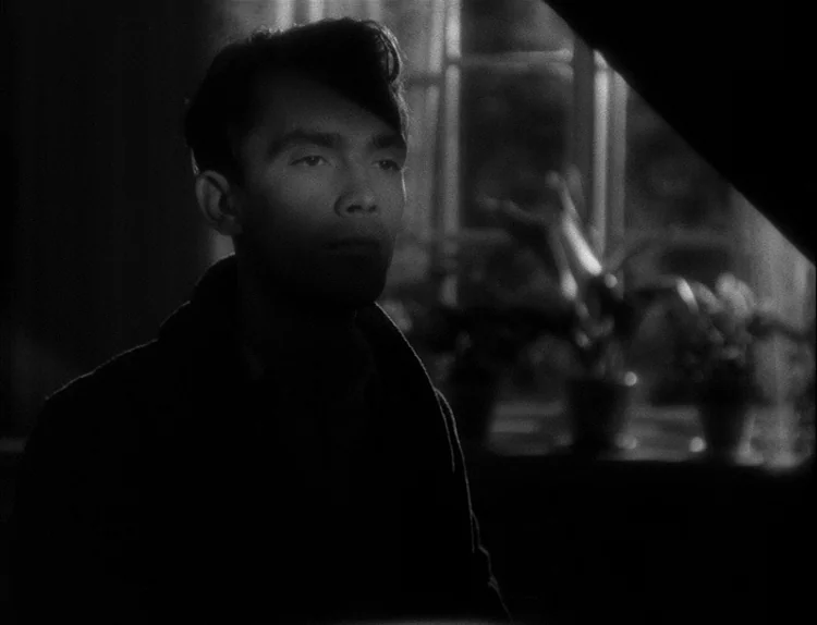 Music in Darkness (1948) movie still: medium — A dimly lit room at night with a window showing soft light behind the subject. A man sits calmly,…